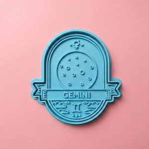 May include: A light blue plastic cookie cutter shaped like an oval plaque featuring the word GEMINI in the center. The design includes a circular pattern of small dots, a planetary symbol at the top, and a Gemini zodiac sign with clouds below.