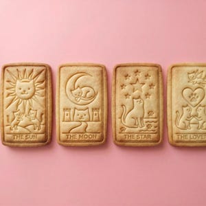 May include: Four rectangular cookies with cat-themed tarot card designs. The cookies are light brown and feature images of the sun, moon, stars, and lovers, each with a cat motif. The text on the cookies reads "THE SUN", "THE MOON", "THE STAR", and "THE LOVERS".