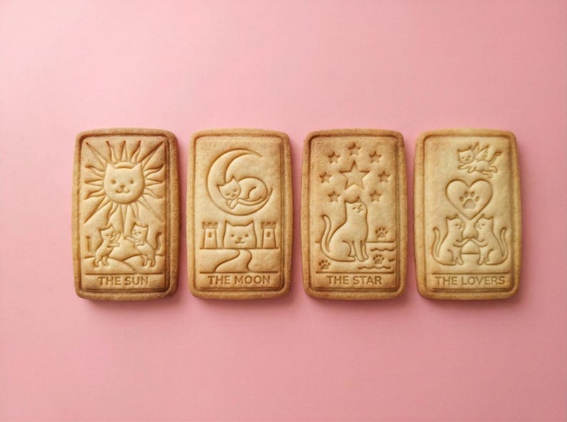 May include: Four rectangular cookies with cat-themed tarot card designs. The cookies are light brown and feature images of the sun, moon, stars, and lovers, each with a cat motif. The text on the cookies reads "THE SUN", "THE MOON", "THE STAR", and "THE LOVERS".