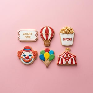 May include: Six decorated sugar cookies featuring carnival themes: a white ticket with the text ADMIT ONE, a red and white hot air balloon, a bucket of popcorn, a clown face, a cluster of colorful balloons, and a red and white striped circus tent.