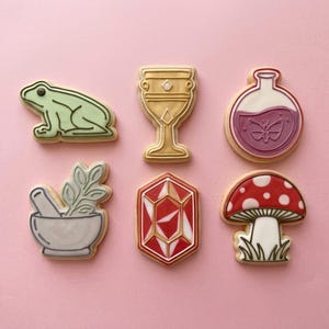 May include: A collection of decorated cookies in a whimsical theme. The cookies include a green frog, a golden chalice, a potion bottle with a butterfly, a mortar and pestle with herbs, a red gem, and a mushroom with white spots.
