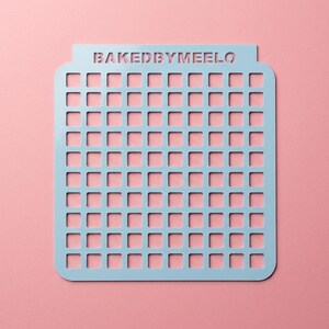 May include: A light blue rectangular stencil featuring a grid of small square cutouts and the text BAKEDBYMEELO laser-cut into the top border. The object has rounded corners and a flat surface, designed for precise patterns on baked goods.