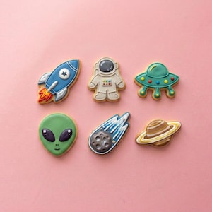 May include: Six space-themed sugar cookies decorated with royal icing. The set includes a blue rocket, a white astronaut, a teal flying saucer, a green alien head, a gray comet with a blue tail, and a brown planet with yellow rings.