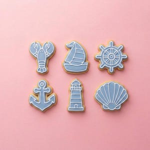 May include: Six nautical-themed cookies on a pink background. The cookies are in the shapes of a lobster, sailboat, ship's wheel, anchor, lighthouse, and seashell. Each cookie has a light blue design with white outlines.