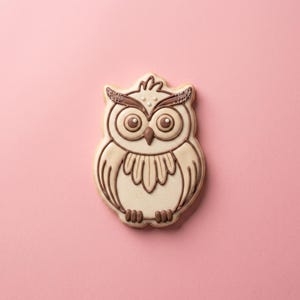 May include: A sugar cookie shaped like an owl, decorated with cream-colored icing and brown piped details. The owl features large round eyes, a pointed beak, and feathered wing patterns, with subtle brown glitter accents on the ear tufts.