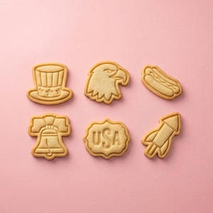 May include: Six light brown sugar cookies arranged in two rows. The shapes include a top hat, an eagle head, a hot dog, a bell, a scalloped plaque with the text USA, and a rocket. Each cookie features embossed details on the surface.