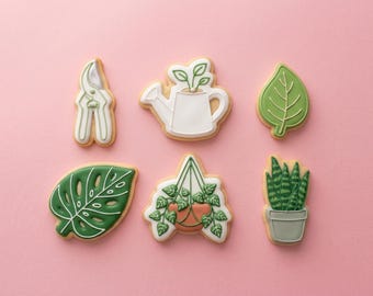 May include: Six sugar cookies decorated with garden-themed icing. The shapes include white pruning shears, a white watering can with a sprout, a green leaf, a dark green monstera leaf, a hanging plant in a terracotta pot, and a snake plant in a pot.