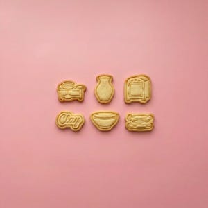 May include: Six light tan cookie cutters arranged in two rows. The top row features a pottery wheel, a vase, and a kiln. The bottom row includes a cookie with the word Clay, a bowl, and a stack of three plates, all with embossed details.