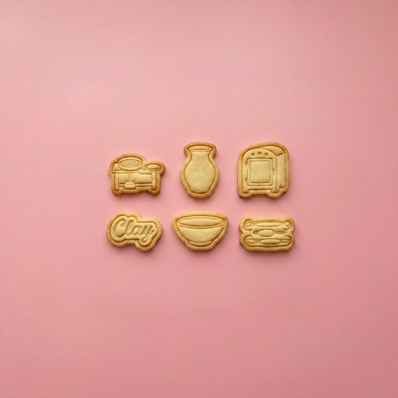 May include: Six light tan cookie cutters arranged in two rows. The top row features a pottery wheel, a vase, and a kiln. The bottom row includes a cookie with the word Clay, a bowl, and a stack of three plates, all with embossed details.