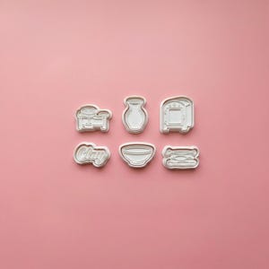 May include: A set of six white plastic cookie cutters featuring pottery-themed shapes including a pottery wheel, a vase, a kiln, the word Clay in cursive script, a bowl, and a collection of sculpting tools arranged in two rows of three.