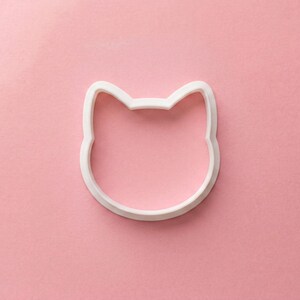 Puede incluir: Un cortador de galletas blanco con forma de cara de gato sobre un fondo rosa. El cortador es de pl&aacute;stico y tiene un dise&ntilde;o sencillo y limpio. Las orejas del gato son puntiagudas y la cara es redonda. El cortador se utiliza para hornear galletas.