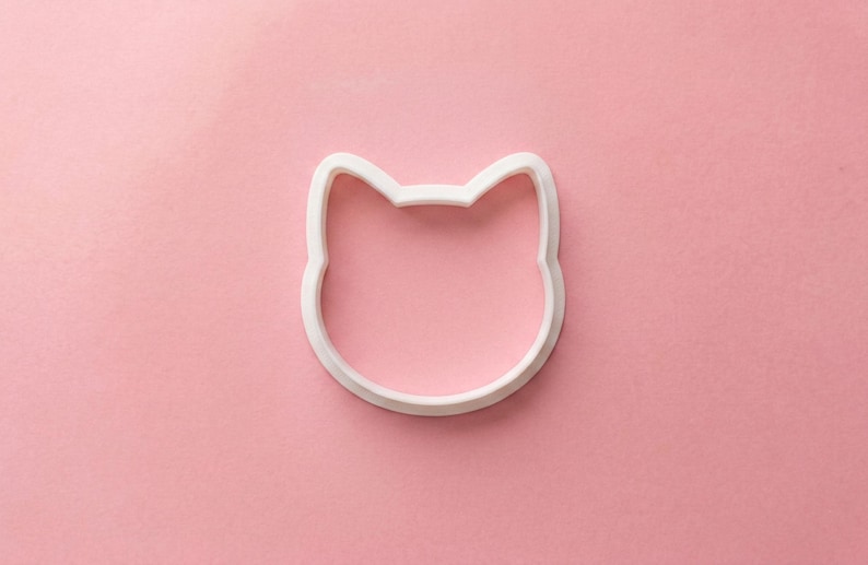 Puede incluir: Un cortador de galletas blanco con forma de cara de gato sobre un fondo rosa. El cortador es de pl&aacute;stico y tiene un dise&ntilde;o sencillo y limpio. Las orejas del gato son puntiagudas y la cara es redonda. El cortador se utiliza para hornear galletas.