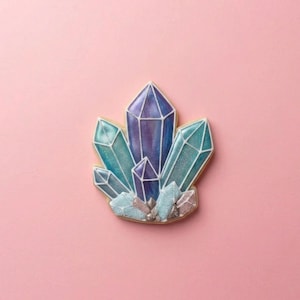 May include: A crystal-shaped cookie with a light brown base, featuring a cluster of blue, teal, and purple crystals outlined in white. The cookie is set against a solid pink background, creating a visually appealing contrast.