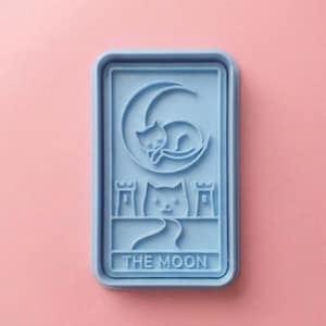 May include: A light blue rectangular mold featuring a cat curled around a crescent moon, with another cat below. The text "THE MOON" is at the bottom. The mold is on a pink background.