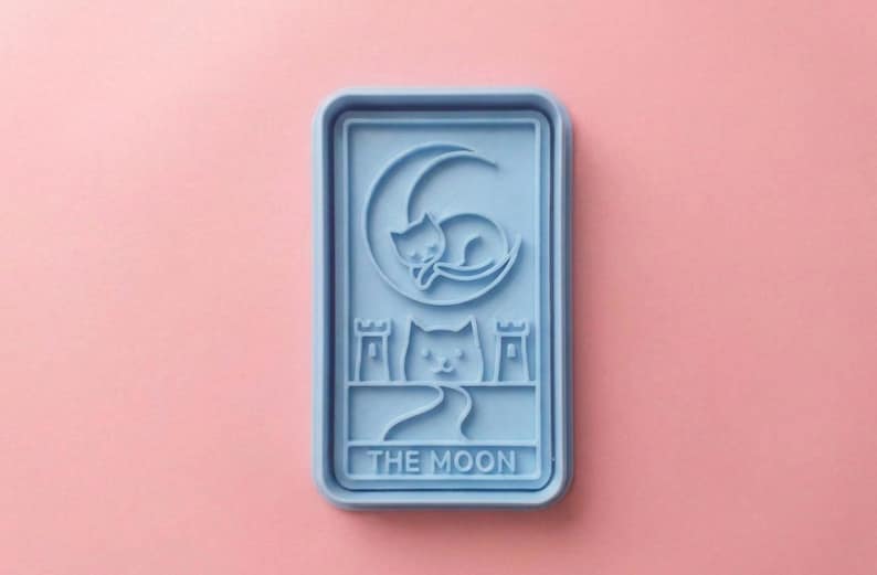 May include: A light blue rectangular mold featuring a cat curled around a crescent moon, with another cat below. The text "THE MOON" is at the bottom. The mold is on a pink background.