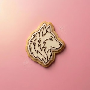 May include: A sugar cookie shaped like a wolf head, featuring a light tan baked edge and a smooth, off-white icing surface. The wolf design is outlined in dark brown icing, detailing the ears, fur texture, snout, and a small dark nose.