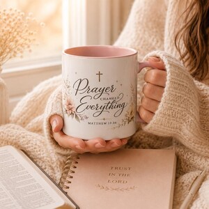 May include: A white ceramic mug with a pink interior, featuring the text "Prayer Changed Everything" and floral accents. The mug is held in a cozy setting with a book, candle, and notebook. The text "Matthew 19:26" is also visible.