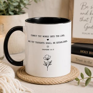 May include: A white ceramic mug with a black interior and handle. The front features black text that reads: COMMIT THY WORKS UNTO THE LORD, AND THY THOUGHTS SHALL BE ESTABLISHED. (PROVERBS 16:3), accompanied by a small heart, a sun icon, and a flower line drawing.