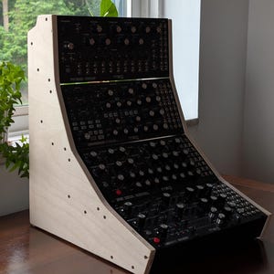 May include: A three-tier Moog synthesizer rack system featuring a light-colored wooden frame with side panels containing circular cutouts. The black synthesizer modules are stacked vertically, each equipped with numerous control knobs, switches, and labels.