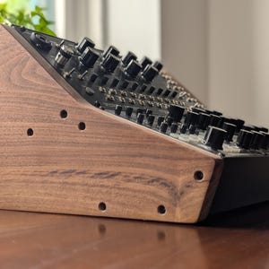 May include: A wooden synthesizer stand with a dark walnut finish. The stand has a slanted design and features several drilled holes. The synthesizer has numerous black knobs and buttons.