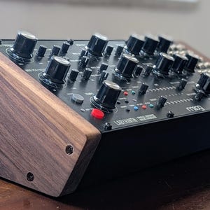 May include: A black synthesizer with numerous knobs and switches, featuring a wooden side panel. The device has a red button and the text "Moog" is visible. The synthesizer is designed for music production.