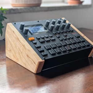 Op de afbeelding: Een zwarte Digitakt II drumcomputer en sampler met een centraal digitaal display, talloze grijze draaiknoppen en een raster van zwarte vierkante knoppen, geplaatst in een op maat gemaakte lichtgekleurde houten standaard.