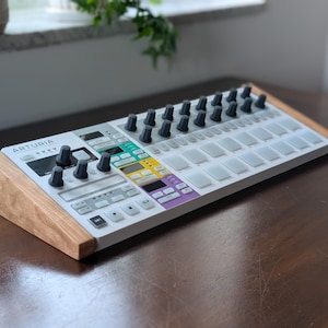 May include: An Arturia MIDI controller keyboard featuring a white chassis with light wood side panels. The interface includes a grid of backlit square pads, a row of black rotary knobs, and a control section with colorful buttons and small screens.
