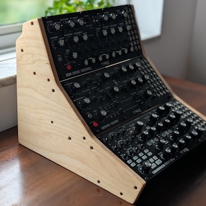 May include: A wooden synthesizer stand with three black synthesizer modules. The stand is made of light-colored wood and has a slanted design. The modules have numerous knobs and buttons. The word "Moog" is visible on the modules.