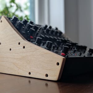 May include: A black electronic music synthesizer with numerous knobs and buttons, mounted on a light-colored wooden stand. The stand has several drilled holes and a slanted design. The synthesizer is positioned on a dark wooden surface.