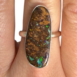 May include: A silver ring featuring a large, oval-shaped opal. The opal has a dark brown base color with vibrant flashes of green, blue, and yellow. The stone is set in a polished silver band.