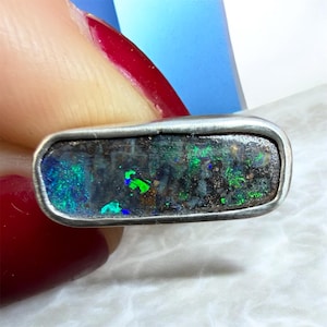 Pode incluir: Um anel retangular com uma pedra escura polida que apresenta flashes iridescentes vibrantes de verde e azul. A pedra está engastada numa moldura de metal prateado liso e escovado que emoldura a forma alongada da gema.