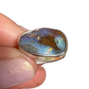 May include: A silver ring featuring a large, oval-shaped opal gemstone. The opal displays a mix of colors, including blue, brown, and hints of green and yellow. The stone is set in a simple, polished silver band.