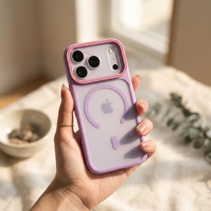 May include: A lavender smartphone case with a translucent back panel, featuring a raised coral-colored camera frame and a circular magnetic charging ring design. The case includes a central Apple logo cutout and a vertical alignment magnet indicator.
