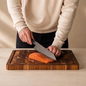 Puede incluir: Una persona corta salmón con un cuchillo grande sobre una tabla de cortar de madera. Un limón a la mitad y un paño beige doblado están sobre la mesa. También hay un pequeño jarrón con plantas secas.