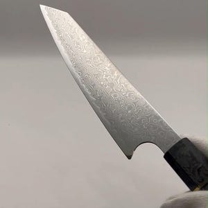Peut inclure: Un couteau de cuisine doté d'une lame pointue et tranchante présentant un motif ondulé complexe en acier damas sur toute la surface métallique. La lame possède une découpe incurvée près de la mitre, rejoignant un manche sombre et texturé.