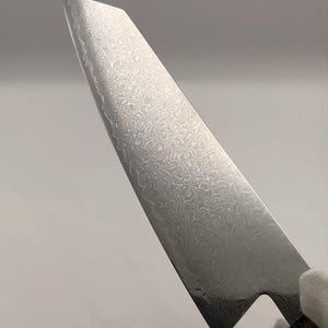 Peut inclure: Un couteau de cuisine avec une pointe acérée et une lame métallique présentant un motif complexe en acier damassé ondulé. La lame possède une finition polie qui reflète la lumière, révélant des lignes tourbillonnantes sur toute sa surface.