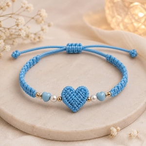 Può includere: Un braccialetto in macramè azzurro con un ciondolo centrale a forma di cuore intrecciato, affiancato da piccole perle bianche e perline dorate. Il braccialetto include due perline aggiuntive a forma di cuore azzurro e una chiusura scorrevole.