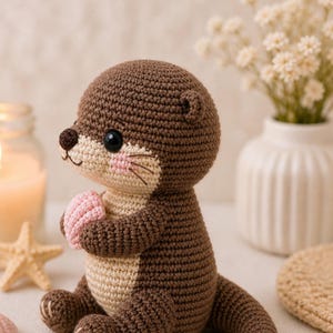 Op de afbeelding: Een gehaakte otter knuffel in bruin, beige en roze tinten. De otter houdt een kleine roze schelp vast. Het heeft zwarte ogen, een beige gezicht en roze wangen. De knuffel zit, met zijn poten zichtbaar.