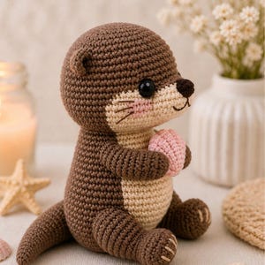 Op de afbeelding: Een gehaakte otter knuffel in bruin- en beigetinten, met een roze bal. De otter heeft een gedetailleerd gezicht met zwarte ogen, een bruine neus en roze wangen. Het handgemaakte speelgoed zit op een wit oppervlak.
