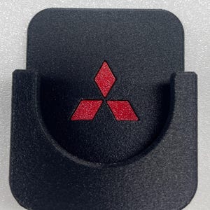 Op de afbeelding: Een zwarte, vierkante houder met een U-vormige uitsparing in het midden. Het oppervlak is voorzien van een verzonken, rood Mitsubishi-logo met drie diamanten. Het object heeft een getextureerde, matte afwerking en een platte onderkant.