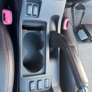 May include: A dark gray vehicle center console featuring two circular cup holders, a handbrake lever with red stitching, two heated seat switches, and an all-wheel drive control switch. The console is positioned between two black seats with red stitching.
