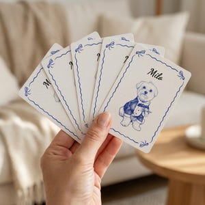 Hand holding a set of custom playing cards featuring a blue hand-drawn line art illustration of a pet in a tuxedo with a decorative bow border and name "Milo".
