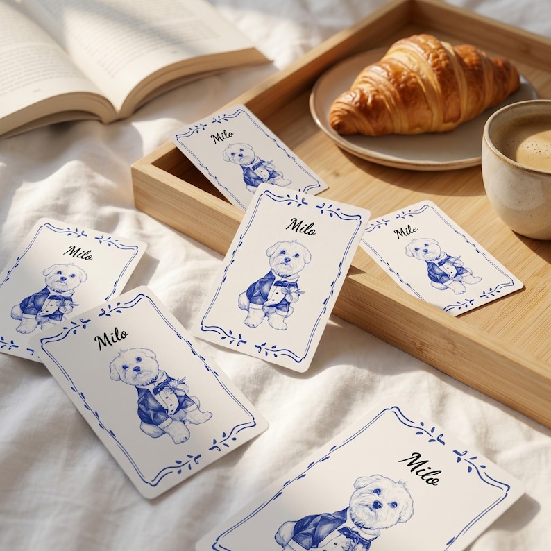 May include: Rectangular cards featuring a blue and white illustration of a small dog wearing a tuxedo, with the name Milo printed above the dog. Each card has a decorative blue border with leaf motifs, presented on a white fabric surface.