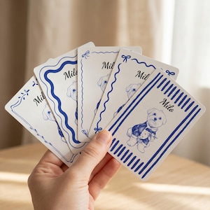 May include: A set of five rectangular white cards fanned out, each featuring a blue ink illustration of a small dog wearing a suit and the name Milo printed in script. The cards have various blue decorative borders including stripes and wavy lines.