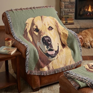 Op de afbeelding: Een groene geweven deken met een afbeelding van een golden retriever. De deken heeft een gefranjerde rand. Een bijpassend sierkussen is zichtbaar. Een mok en een boek met de titel "The Good Dog" staan op een bijzettafel.