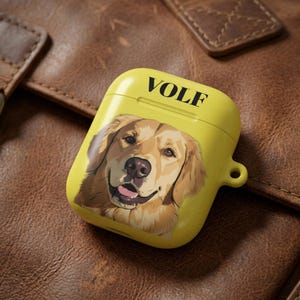 May include: Yellow AirPod case featuring a Golden Retriever portrait. The case has the word "VOLF" printed in black at the top. The case is resting on a brown leather surface.