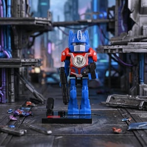 May include: A blue, red, and white toy robot figure with two black guns. The robot has a blue head with antenna-like features and a red chest with a circular emblem. It stands on a black base with the word "TRANSFORMERS" printed on it.