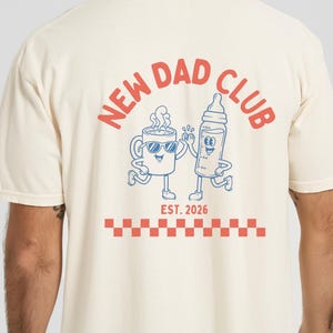May include: Cream-colored t-shirt with "NEW DAD CLUB" text in red on the front and back. The back features a cartoon coffee cup and baby bottle, with "EST. 2026" below. A red and white checkered pattern is at the bottom.