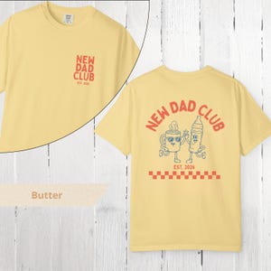 May include: Butter-colored t-shirt with "NEW DAD CLUB" text and graphic on the front and back. The back graphic features a coffee cup and baby bottle with sunglasses, and the text "EST. 2026".
