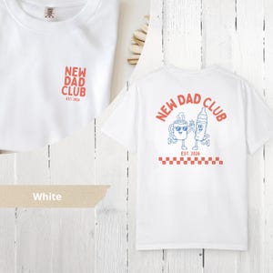 May include: White t-shirt with "NEW DAD CLUB" in red text on the front and back. The back features a cartoon coffee cup and baby bottle with sunglasses, and the text "EST. 2026". A red and white checkered pattern is at the bottom.